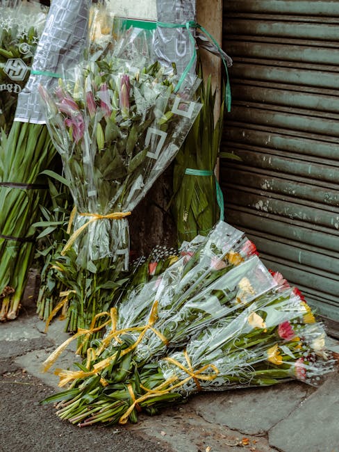 Multiple bouquets of fresh flowers resting against a weathered wall and closed metal shutter on a sidewalk. The bouquets include pink lilies and roses, wrapped in clear cellophane tied with yellow rib