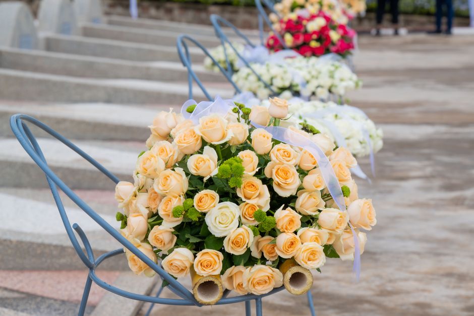 A floral arrangement of fresh peach and cream roses, accented with small green pom-pom chrysanthemums and wrapped with white ribbon, placed within a blue metal chair on a stone-paved outdoor setting. 