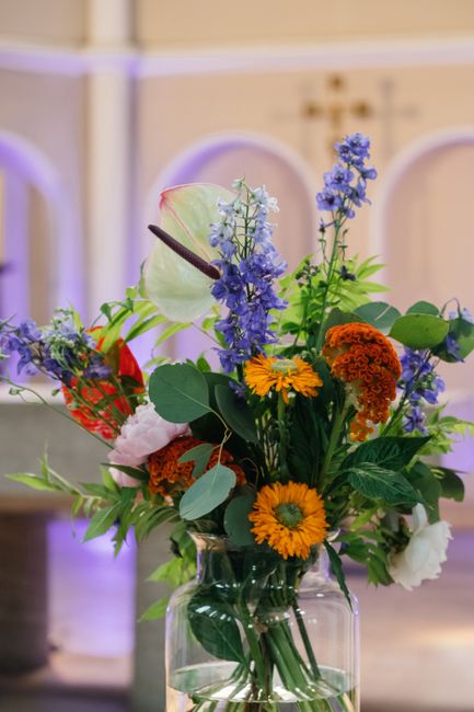A vibrant floral arrangement in a clear glass vase featuring a variety of fresh flowers, including bright yellow daisies, orange marigolds, and delicate purple delphiniums. The bouquet is accented wit