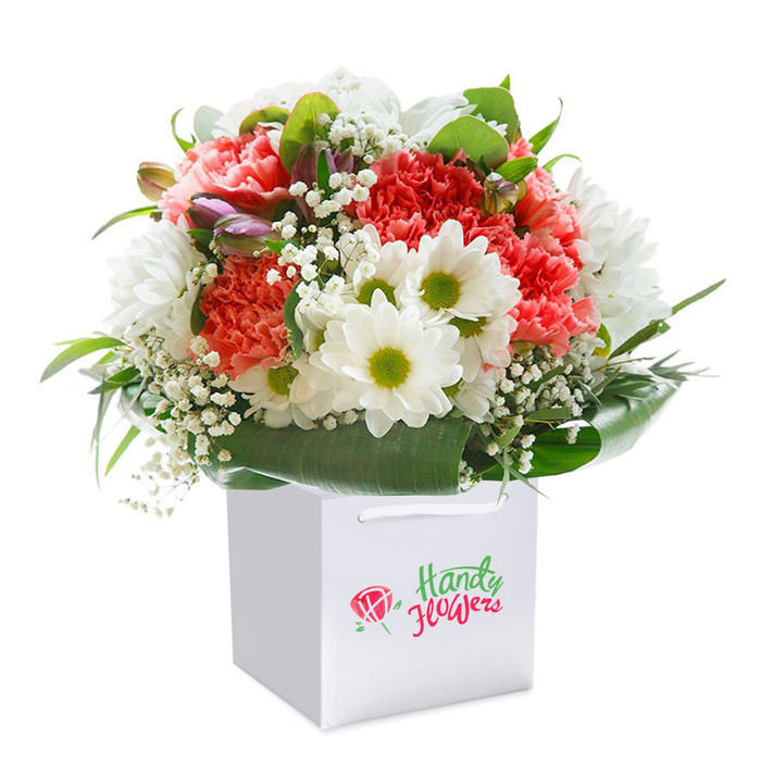Nestled within the clean lines of a white square gift bag, this stunning bouquet captures the lively essence of Tooting Bec with a masterful blend of colour and form. A wealth of lush carnations in coral and warm peach hues take centre stage, each bloom unfurling in a profusion of soft, frilled petals. Interwoven among these are classic white daisies, their satiny petals outlining luminous yellow-green centres that bring a flash of cheerful brightness, as vibrant as the neighbourhood's bustling street markets. Baby's breath (gypsophila), with its tiny, ethereal white blossoms, is delicately interspersed to soften and lighten the overall feel, fluttering between the larger flowers like whispers of spring air drifting through Tooting Bec Common. Subtle purple-tinged buds nestle quietly amid the arrangement, lending subtle depth and gentle intrigue to the left side. All is surrounded by lush deep green foliage-broad, glossy leaves arch elegantly to frame the bouquet, offering a natural structure and a sense of gathered, organic beauty. The bouquet's composition achieves a pleasing rounded silhouette, imbued with freshness and movement. The white bag, complete with robust ribbon handles, stands ready for delivery or a joyful presentation at a friend's home in SW17. Photographed against an immaculate white background, the arrangement radiates sophistication and care, a true testament to the skill and attention of local Tooting Bec florists, ideal for anything from milestone celebrations to simple acts of kindness.